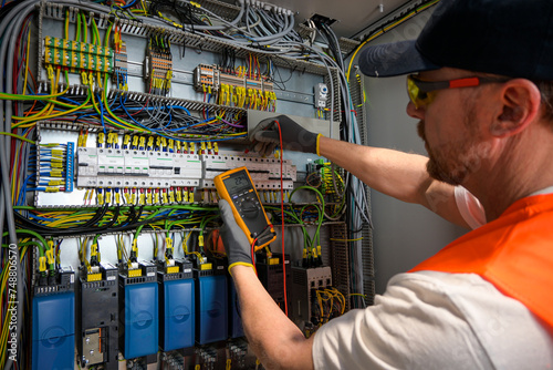 an electrician in a cap, yellow glasses and an orange vest measures electric current with a digital multimeter on a distribution box