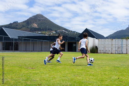 Two boys are playing soccer on a grassy field with mountains in the background in school