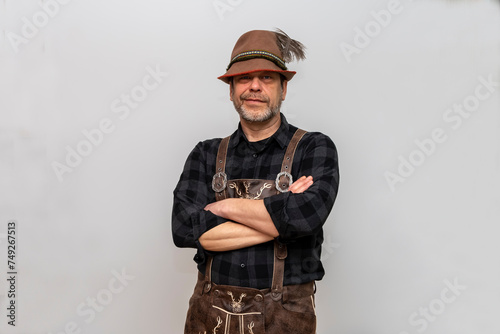 Studio portrait of a man wearing a Tyrolean feather hat and leather shorts, arms crossed on his chest, white background.