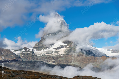 Low angle view of near symmetric pyramidal peak of the Matterhorn near Zermatt, Switzerland in extended Monte Rosa area of the Pennine Alps somewhat surrounded by white clouds in otherwise blue sky