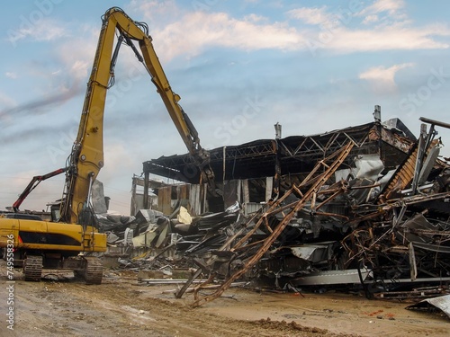 Excavator dismantling the charred remains of a burnt warehouse, twisted metal structures visible