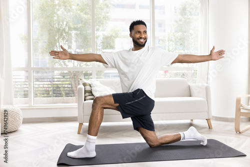 Happy athletic young African man exercising at home, training body on yoga mat, enjoying fitness activity, doing twisting lunges, stretching hips, building strong core muscles, looking away, smiling