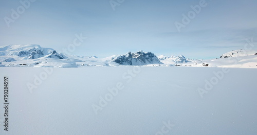 Winter Antarctica snow covered nature landscape. Frozen ocean, mountain range in background, sunny weather, blue sky. Desert white land of ice. Polar arctic travel scene. Low angle drone flight