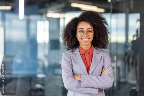 Smiling young African American businesswoman, owner standing in business suit and glasses in office, crossed arms on chest and looking confidently at camera.