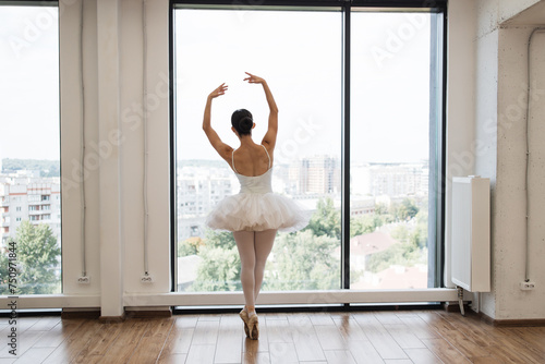 Back view of focused young Caucasian ballerina dressed in white tutu costume. Beautiful female ballet dancer makes pirouette at ballet studio in front of big panoramic window background.