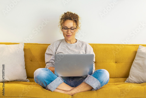 One woman smiling and using laptop computer at home sitting comfortably on a yellow sofa in living room. Smart working female people notebook. Surfing the web. Enjoying technology and connection