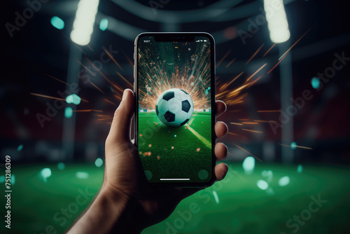 Hand holding a smartphone displaying a soccer ball with spark effects at a stadium.