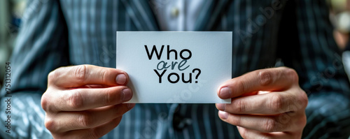 Close up of a man in a suit holding a card with Who Are You? printed on it, questioning identity and self perception