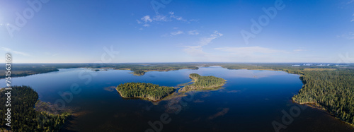 Aerial panoramic view of a northern wilderness lake with small islands. The water is calm and reflecting the blue sky. The surrounding forest is a mix of spruce, pine, and aspen trees. 