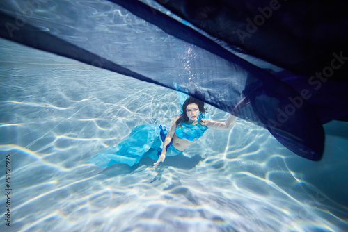 Young black-haired woman in blue dress poses underwater under dark-blue fabric with her eyes open