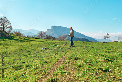 Gray haired man taking a walk with his jack russell dog in meadow in mountainous area. Mature man spending time outdoors with his dog in nature.