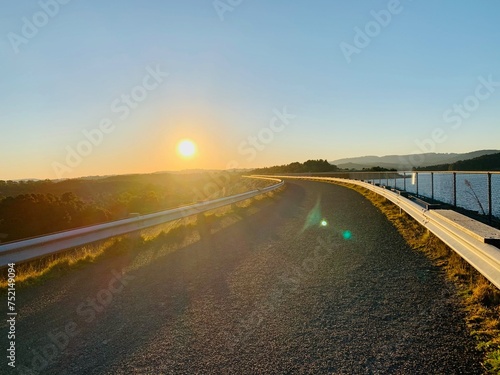 A road with a railing alongside a beautiful forest field and a serene waterscape during sunset