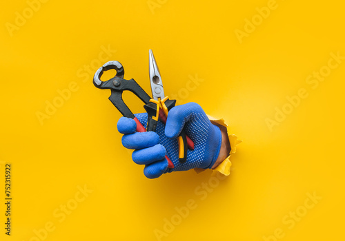 A man's hand in a blue fabric knitted glove holds wire cutters and pliers. Torn hole in yellow paper. The concept of a worker, a labor migrant, a master of his craft. Copy space.