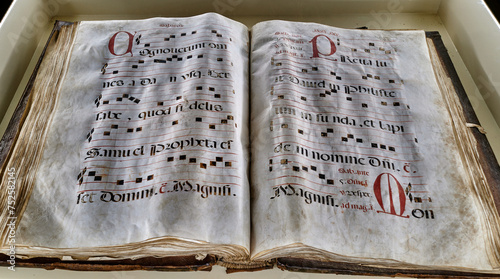 An open, aged book displaying musical notes and lyrics in the style of Gregorian chant.