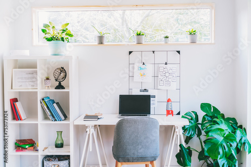 Modern cozy light workplace - white desk with laptop black screen, grid mood board with pinned notes, shelves with docs and green monstera plant at work space in home office room interior