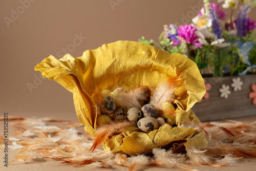 Crepe paper nest with easter eggs, feathers, and spring flowers on a beige background.