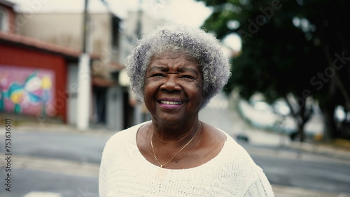One mature black senior woman walking in city street outside. African American elderly lady in 80s strolling in urban environment looking at camera