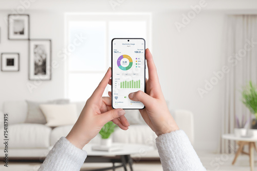 Woman holds smartphone with electricity usage app, monitoring and analyzing consumption for smart home. Living room background. Concept of energy efficiency