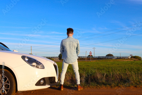 Curly haired Caucasian man peeing on a road with the car next to it. You can see the road and the sky in the background. Concept of stop to take a pee