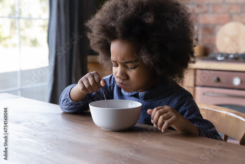 Little 6s cute African curly-haired girl sits at dining table in kitchen frowning while eats not delicious breakfast. Lack poor of appetite, healthy but not tasty monotonous food, not hungry concept