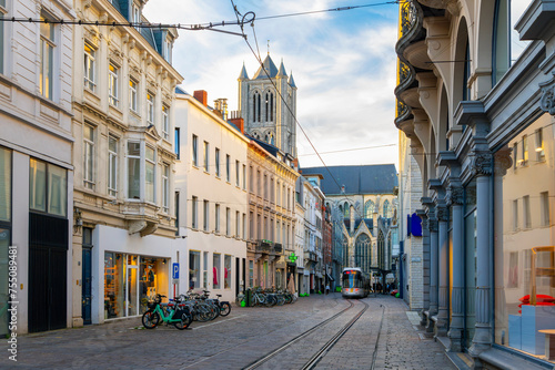 A tram on tracks comes through Sint-Niklaasstraat, or Saint Nicholas Street, with the tower and nave of the Saint Nicholas Cathedral in view behind, in Ghent.