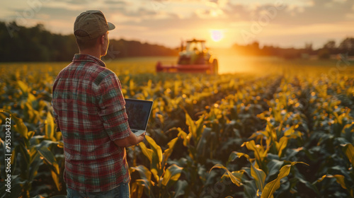 Asian young farmer with laptop standing in corn field, tractor and combine harvester working in corn field in background.