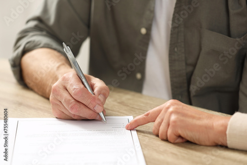 Notary pointing to senior man where to sign Last Will and Testament at wooden table indoors, closeup