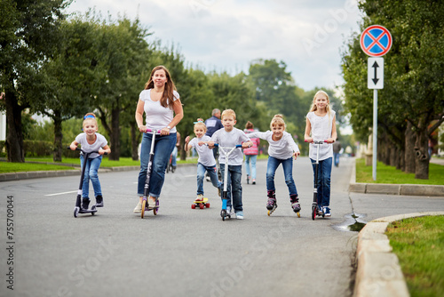 Woman and five children on scooters, roller skates and skateboard ride on street