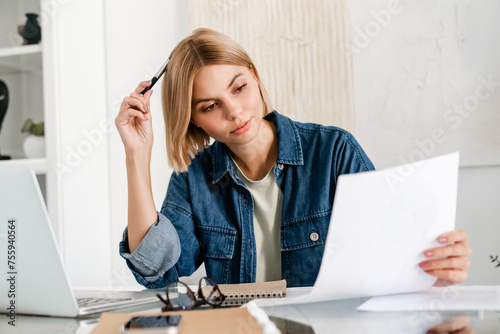 Pensive freelancer doing paperwork working on the laptop. Caucasian young woman holding report, mortgage document, project, solving dealing with problem