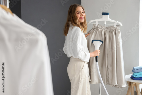 Pretty young woman steaming clothes with modern garment steamer in laundry room