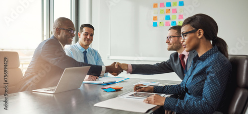 Two diverse business teams sit at a conference table and shake hands on reaching an agreement. They all smile at having made a business deal