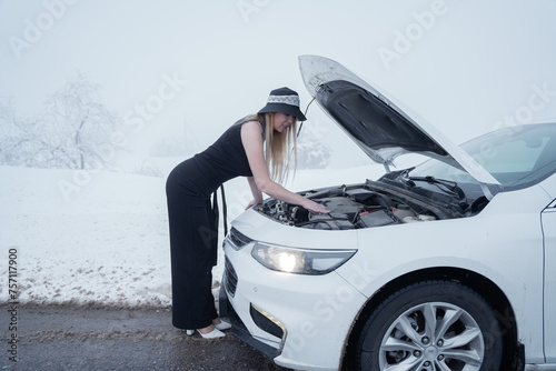 Woman in winter clothing checking under car hood on a snowy road