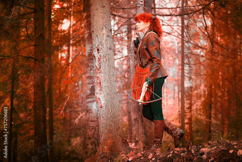 Mature model portraying a royal huntress with red curve hair is hunting with a crossbow in the in vibrant autumn forest in a thematic photo shoot