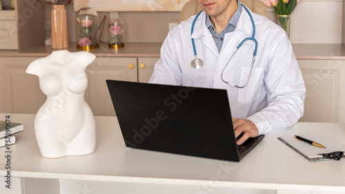 Male general practitioner taking notes in medical history of patient, writing down symptoms, complaints and anamnesis. Handsome doctor in white uniform with stethoscope working on laptop in the office