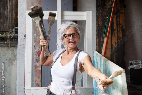 smiling laughing happy older mature woman portrait, proud artist, in her fifties with grey hair and black glasses, with a positive gesture persuasiveness and charisma and many paintbrushes