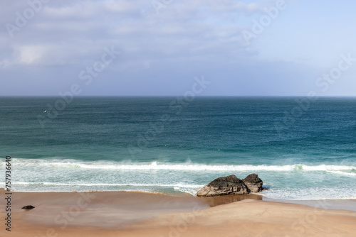 A serene view of Durness Beach, showcasing the tranquil blue waters of the Atlantic meeting the golden sands. A solitary rock formation stands near the shoreline, under a vast, partly cloudy sky