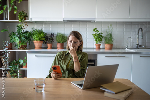 Saddened stressed female sitting at home table with laptop reading disturbing news on mobile phone. Worried nervous woman frustration sadly looking on screen smartphone with negative information.