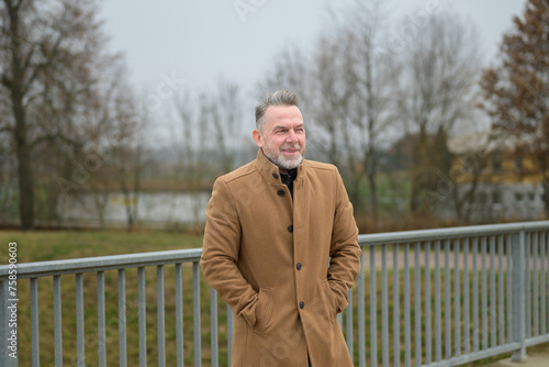 Sideview of amiddle aged man with gray hair and gray beard on a bridge in winter or spring with his hands in pockets