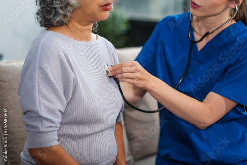 Caucasian female doctor uses stethoscope to check heart rate on elderly Asian patient on sofa.