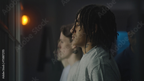 African American teenager stands near prison cell with Caucasian boys, looks at the camera. Young prisoner serves imprisonment term for crime in jail. Youth detention center or correctional facility.