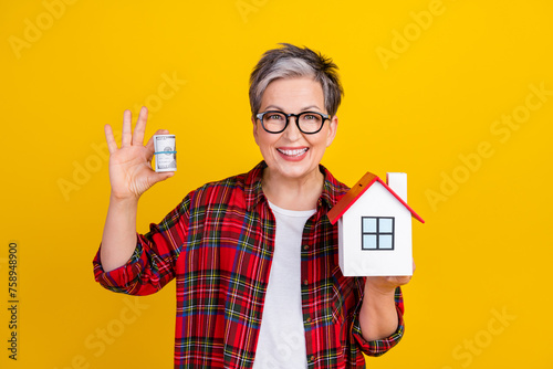Photo of cheerful grandmother hold rolled stack dollars little house isolated on yellow color background