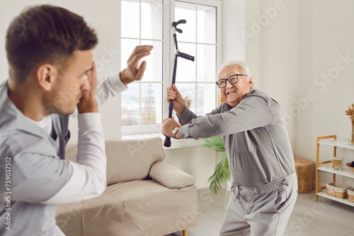 Angry elderly senior man going to hit young male caregiver or nurse with his crutch. Enraged exasperated male pensioner standing at home and threatening scared medical caretaker with cane.