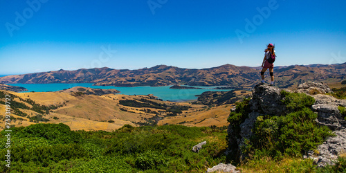 woman with a backpack enjoys the panorama of akaroa harbour from a hill in otepatotu nature reserve, banks peninsula near christchurch, new zealand south island