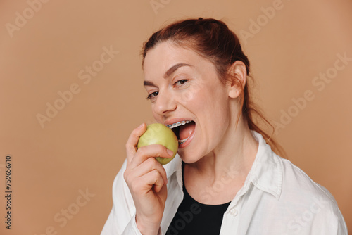 Portrait of mature woman with braces on teeth eating green apple on beige background