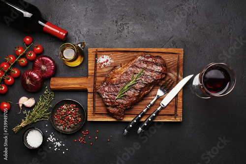 Grilled Striploin beef steak on wooden board with bottle and glass of red wine, vegetables, herbs and spices over black concrete background. Top view