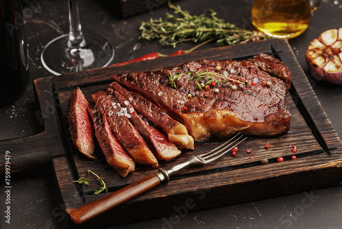 Medium rare sliced grilled striploin beef steak served on wooden board with vintage fork, glass of wine, herbs and spices. Side view, close up
