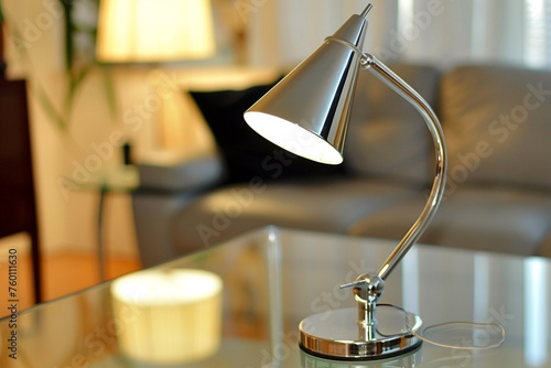 A sleek, chrome desk lamp with a conical shade, illuminating a contemporary glass table in a softly lit room.