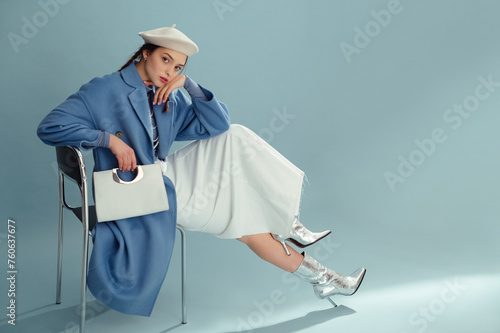 Fashionable confident woman wearing beret, elegant blue coat, white denim maxi skirt, silver ankle boots, holding stylish faux leather purse, posing on blue background. Full-length studio portrait