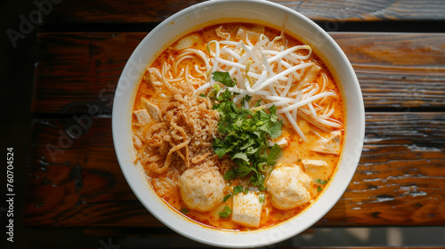 A bowl of laksa with rice noodles, chicken, tofu, bean sprouts, and fish balls, in a spicy coconut curry soup, topped with fried shallots and cilantro.