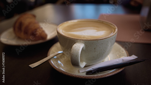 Delicious hot Italian cappuccino stands in a cup with a sugar pa kets on a wooden table in cafe. In the background a fresh croissant on a plate